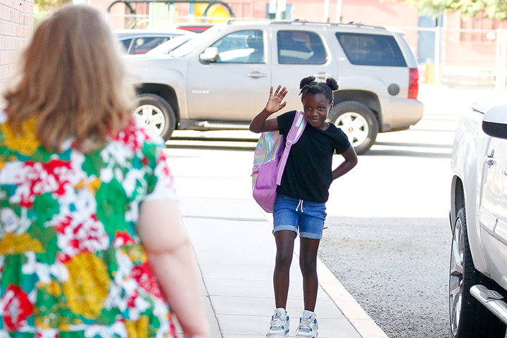 A girl in a black shirt and denim shorts and a purple backpack waves to a woman in a floral dress