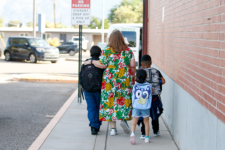 A woman in a floral dress walks with three students next to the school