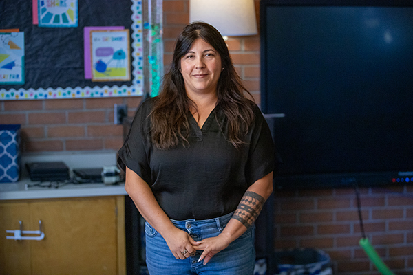 A woman in a black shirt and jeans smiles in her classroom